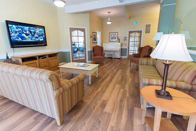 Bright senior living lounge with striped sofas, a coffee table, wall-mounted TV, and armchairs near a fireplace.