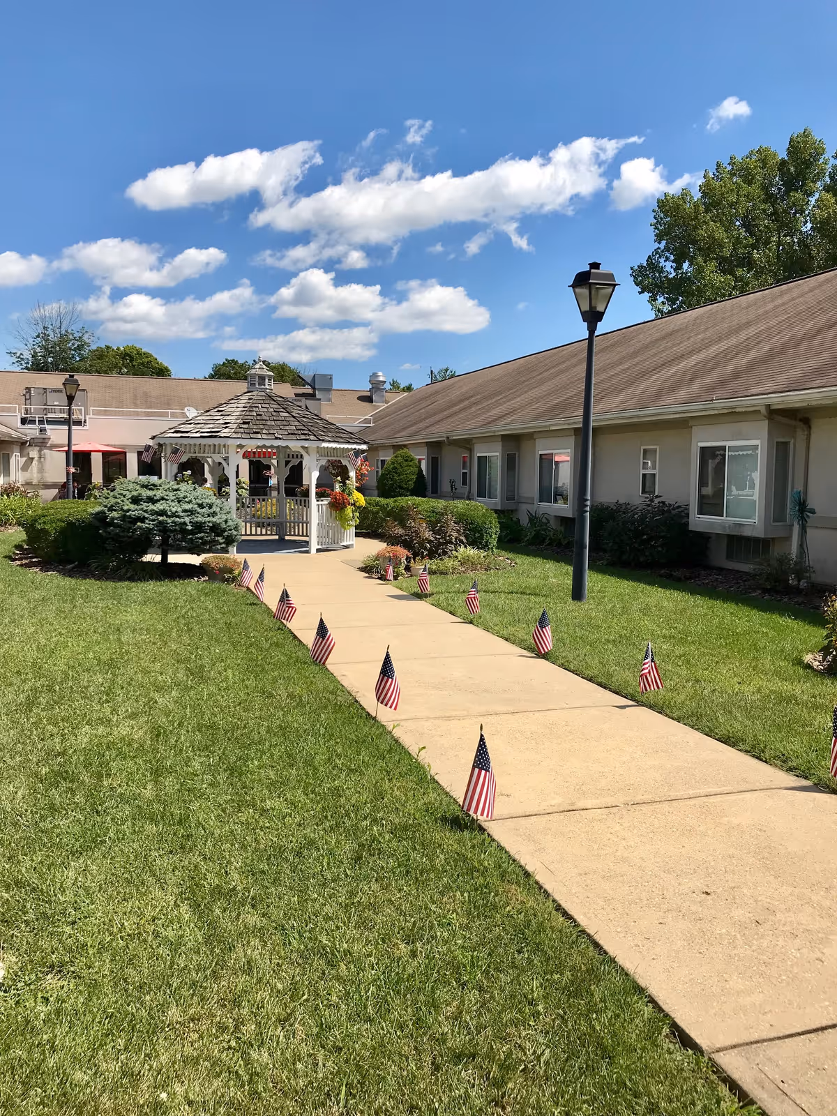 A concrete walkway lined with small American flags leads to a white gazebo in the grassy courtyard of a single-story senior living building.