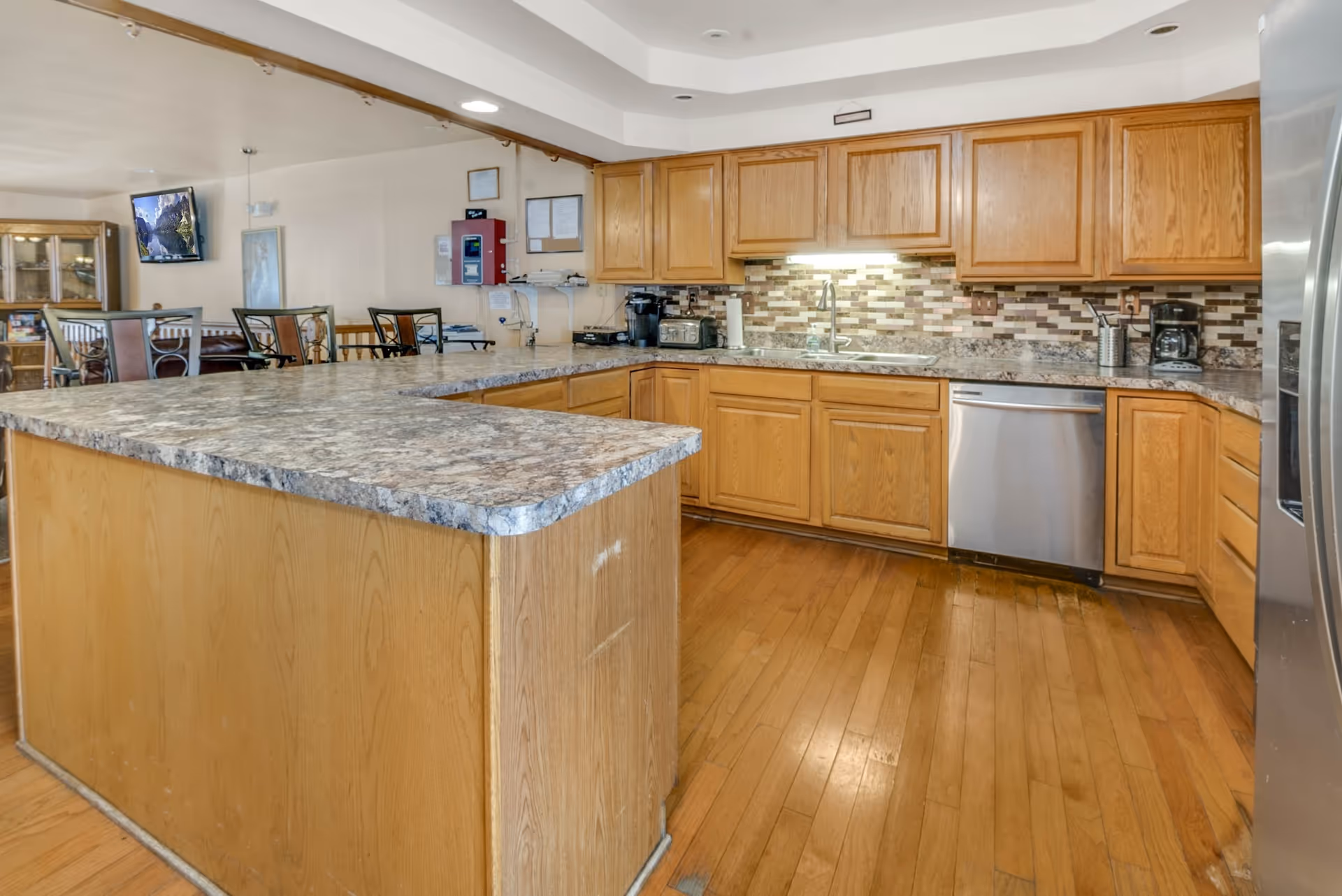 A spacious kitchen area with wooden cabinets, a large granite countertop island, stainless steel dishwasher and refrigerator, and a tiled backsplash. In the background, there is a dining area with chairs and a mounted television on the wall.