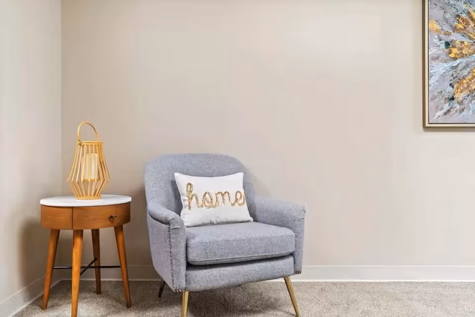 Gray upholstered armchair with a 'home' pillow beside a wooden side table and lamp in a neutral-toned room.