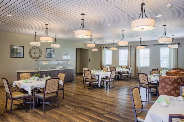 A bright dining room in a senior living facility with several tables covered with white tablecloths, each set with cups, napkins, and small plants. The room has wooden flooring, beige walls, multiple hanging light fixtures, and windows with curtains allowing natural light to enter. There is a counter with a glass sneeze guard along one wall and framed artwork and a large clock on the wall.