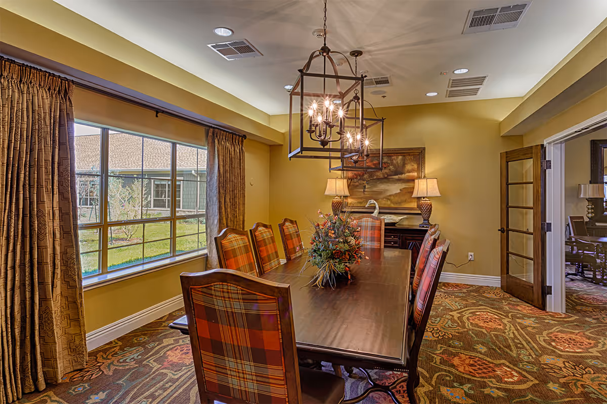 A dining room with a long wooden table, plaid-upholstered chairs, chandelier lighting, and a large window with curtains.