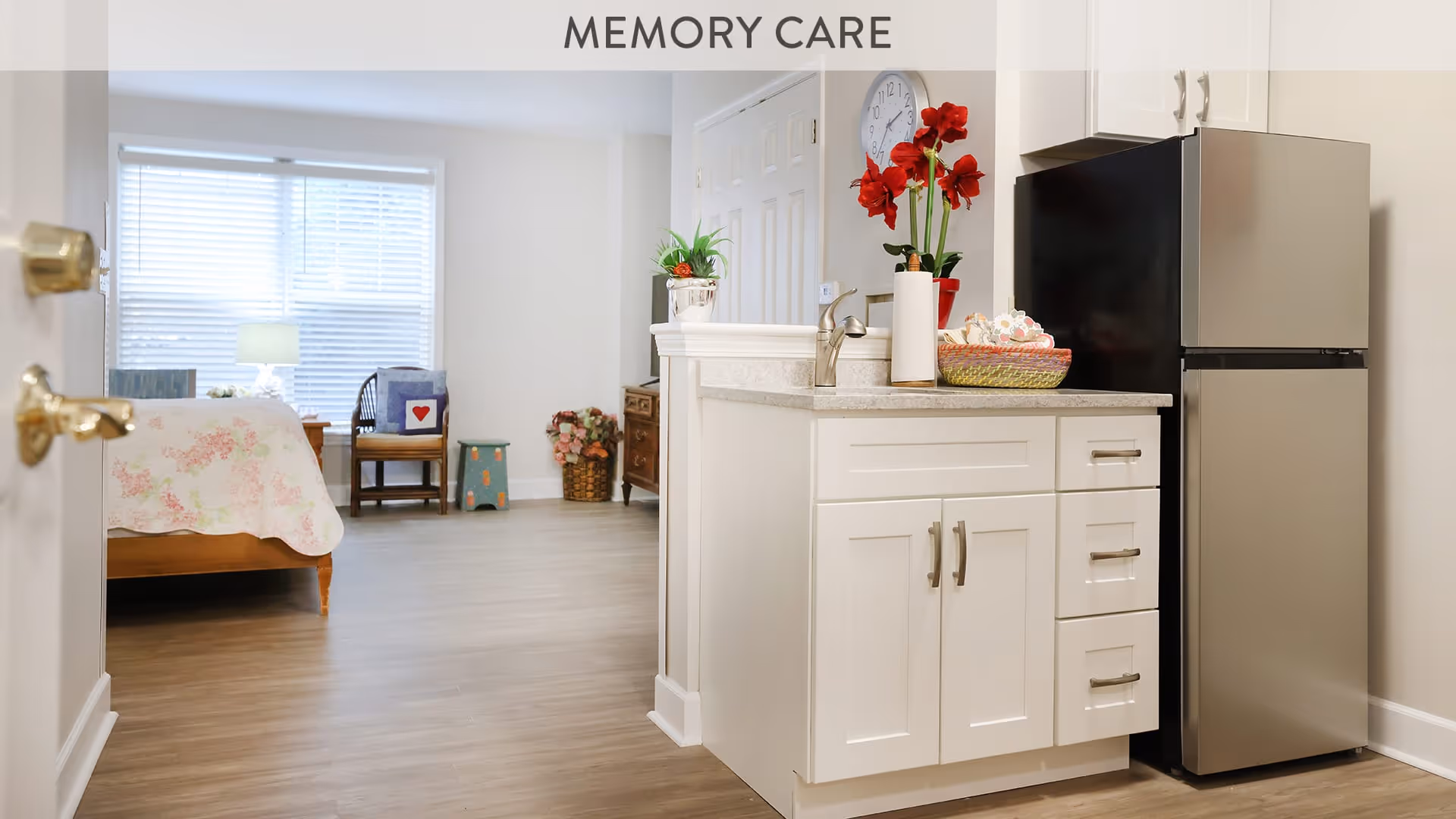 Interior view of a memory care facility showing a small kitchen area with white cabinets, a countertop with a sink, a stainless steel refrigerator, and decorative red flowers. In the background, there is a bedroom area with a bed covered in a floral quilt, a chair with a cushion, a lamp, and a window with blinds letting in natural light.