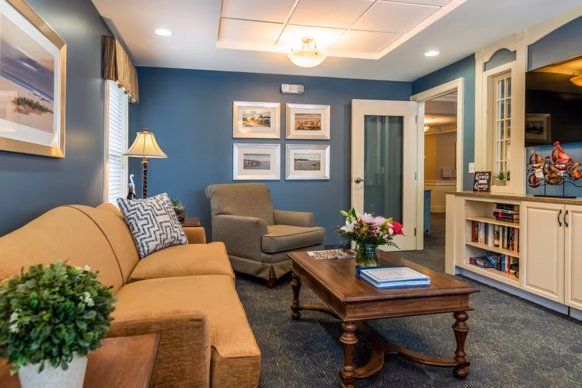 A cozy living room with a mustard yellow sofa, a gray armchair, and a wooden coffee table with a vase of flowers and books. The walls are painted blue and decorated with framed pictures. There is a lamp on a side table next to the sofa and built-in white cabinetry with shelves holding books and decorative items.
