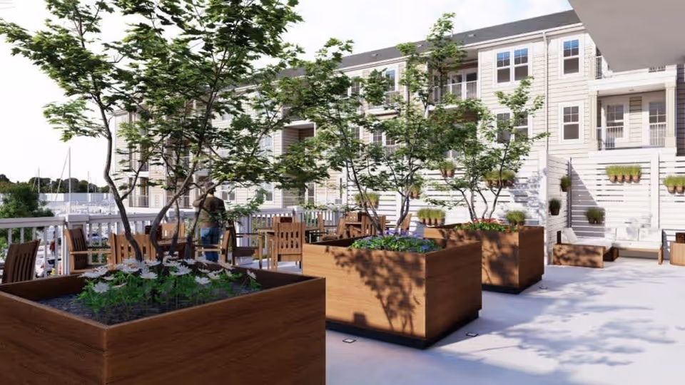 Outdoor patio area with large wooden planters containing small trees and flowers, surrounded by wooden chairs and tables. In the background, there is a multi-story building with balconies and windows, and a marina with boats is visible beyond the railing.
