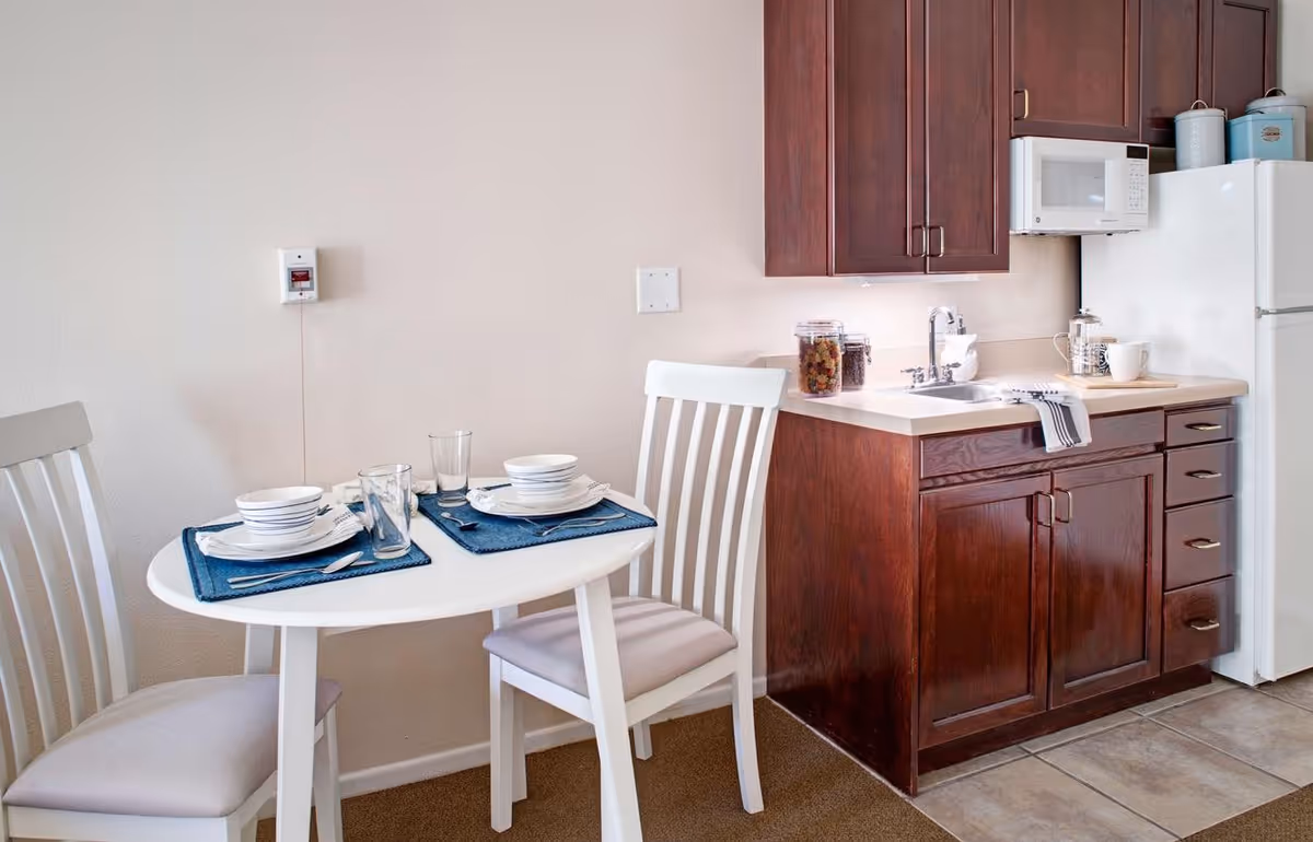Small kitchen area with dark wood cabinets, a white refrigerator and microwave, a sink, and a round table set for two with white chairs.