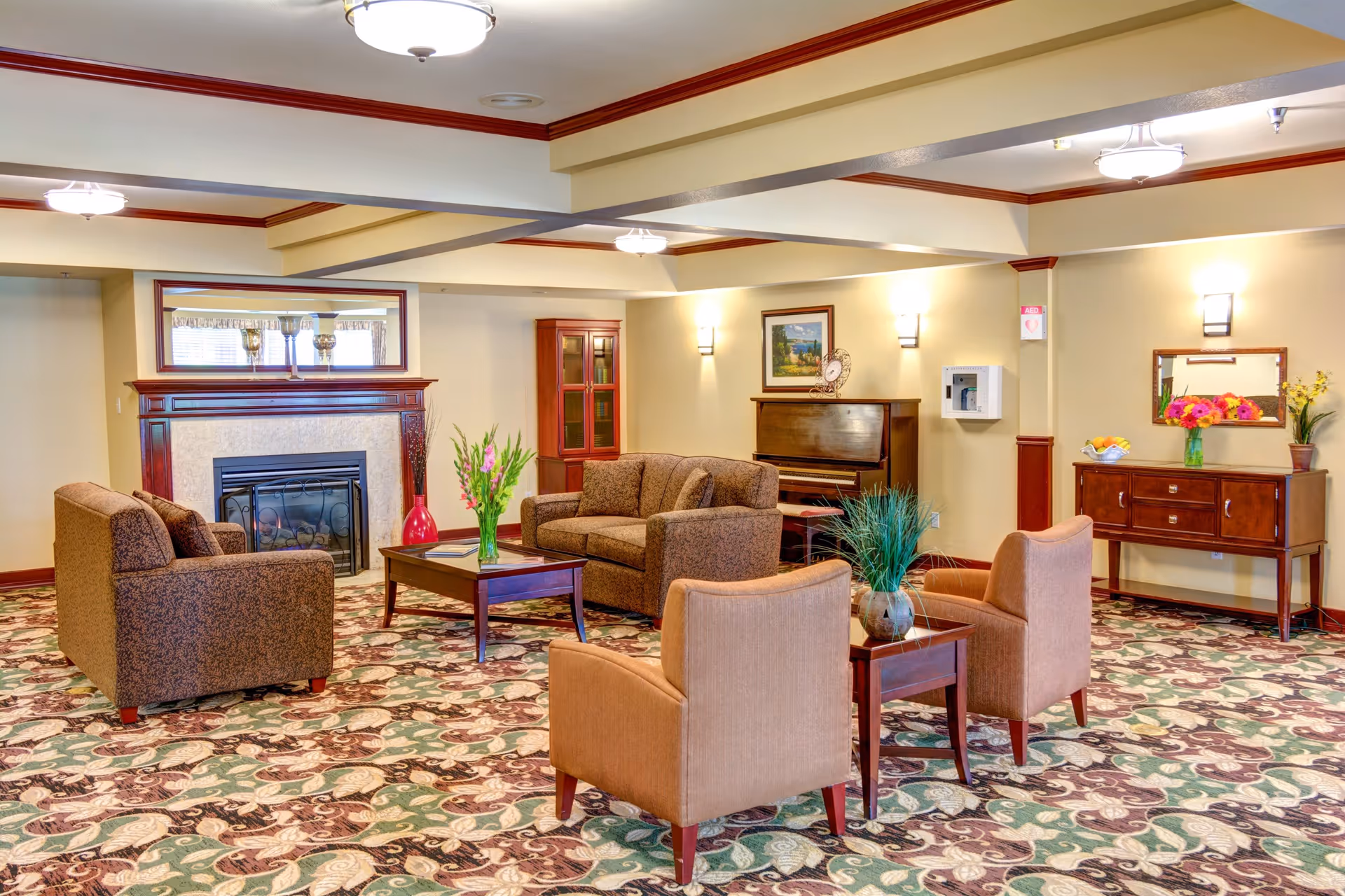 A cozy living room area with patterned carpet, two brown upholstered sofas, two beige armchairs, a wooden coffee table with a vase of flowers, a fireplace with a large mirror above it, a piano against the wall, and a wooden sideboard with a mirror and flower arrangements.
