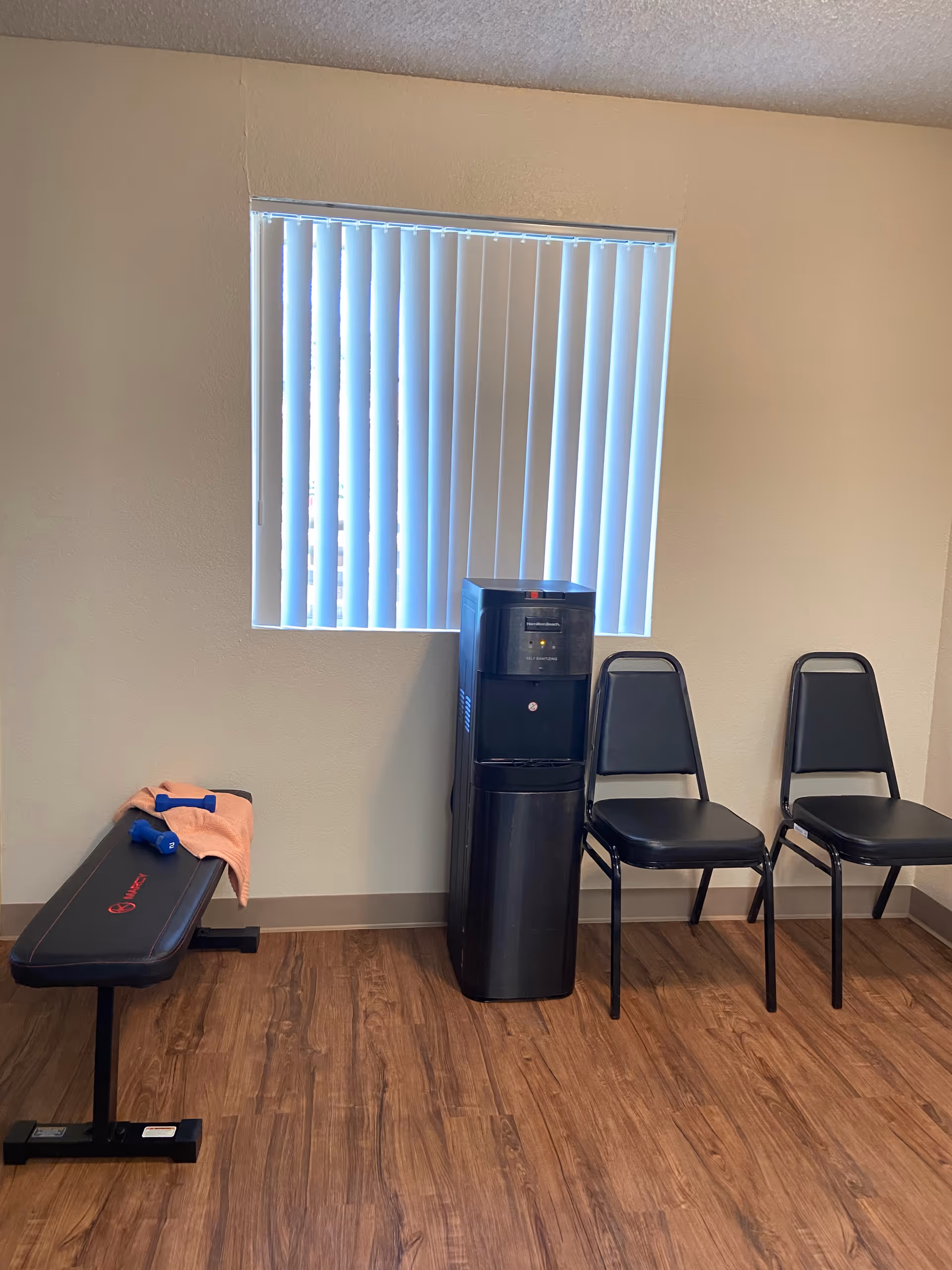A small room with a wooden floor, a window with vertical blinds, a black water cooler, two black chairs, and a black exercise bench with a towel and two blue dumbbells on it.