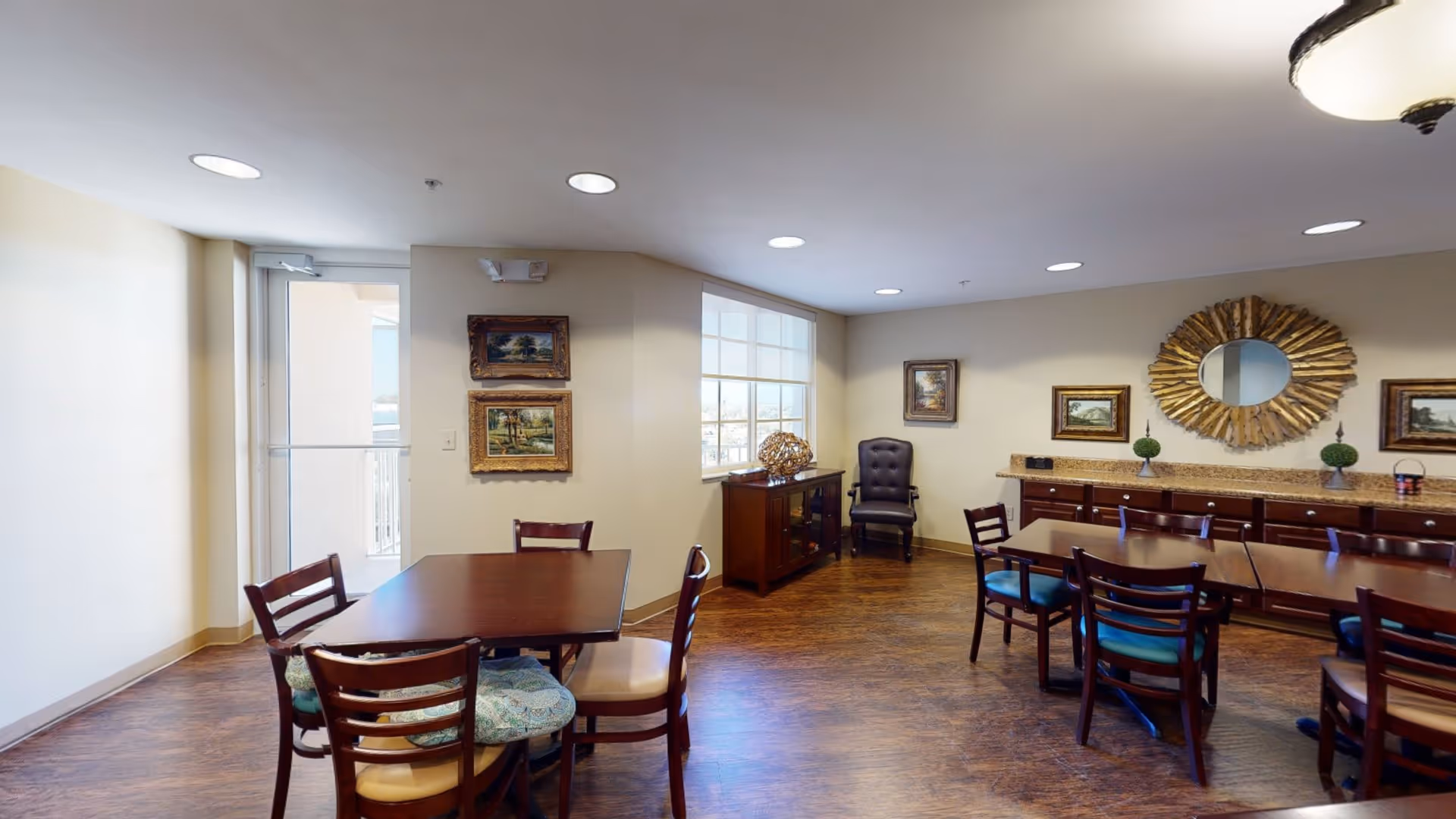 A well-lit dining room with wooden tables and chairs arranged neatly. The room features hardwood flooring, framed landscape paintings on the walls, a large decorative mirror, and a sideboard with decorative items. There is a window allowing natural light and a door leading to an outdoor area.