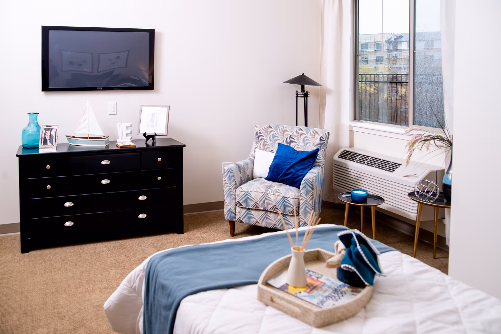 A cozy bedroom featuring a bed with a white quilt and blue throw, a black dresser with decorative items including a photo frame, a model sailboat, and a blue vase. A patterned armchair with blue and white cushions sits next to a window with an air conditioning unit below it. Two small round side tables hold a blue candle and a decorative metal orb. A flat-screen TV is mounted on the wall above the dresser.