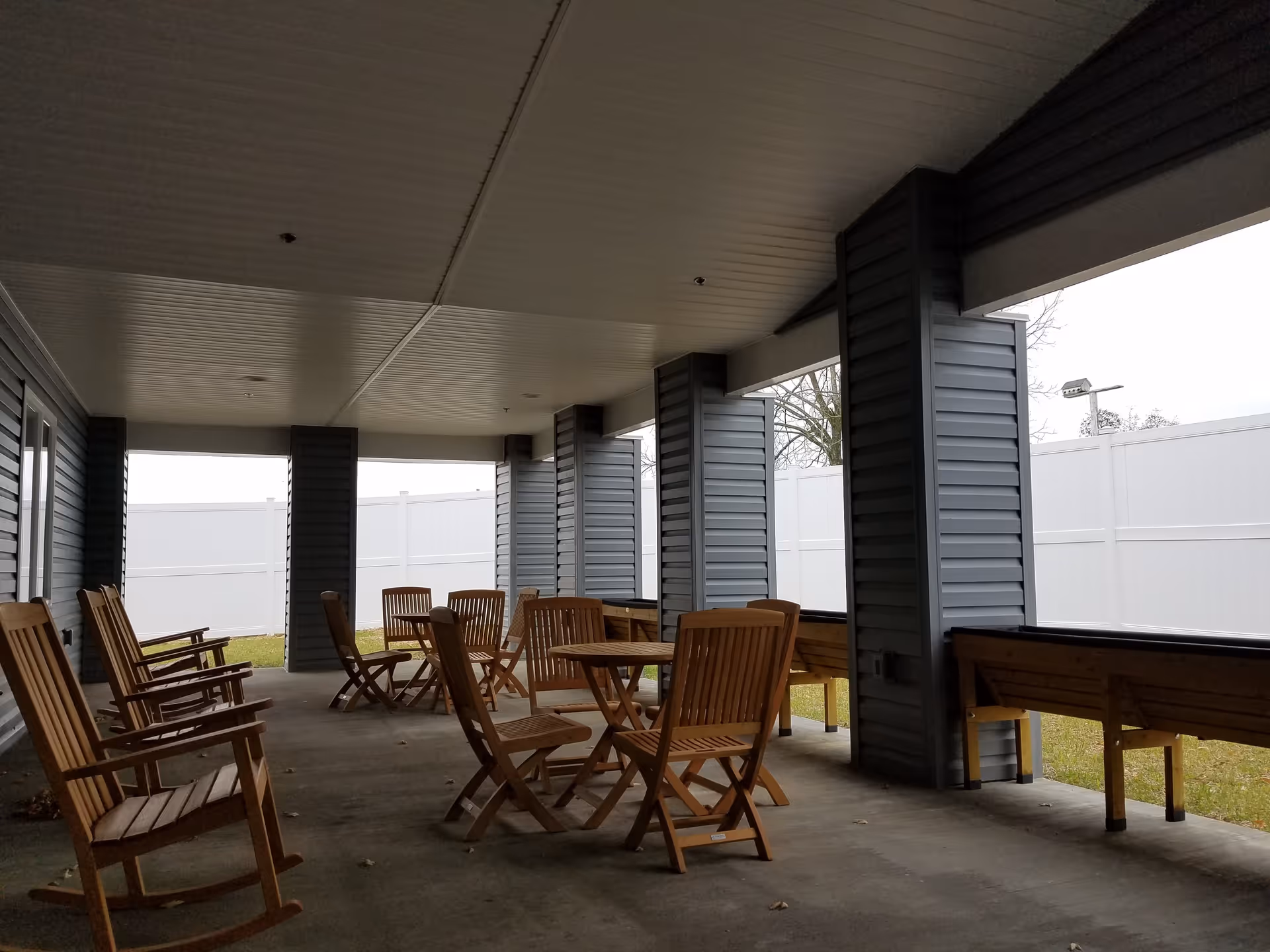 Covered outdoor patio with wooden rocking chairs and tables under a roofed walkway and a white privacy fence.