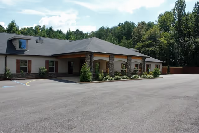Exterior view of a single-story assisted living facility building with a covered entrance supported by stone pillars, surrounded by greenery and trees under a partly cloudy sky. The parking lot in front is empty.