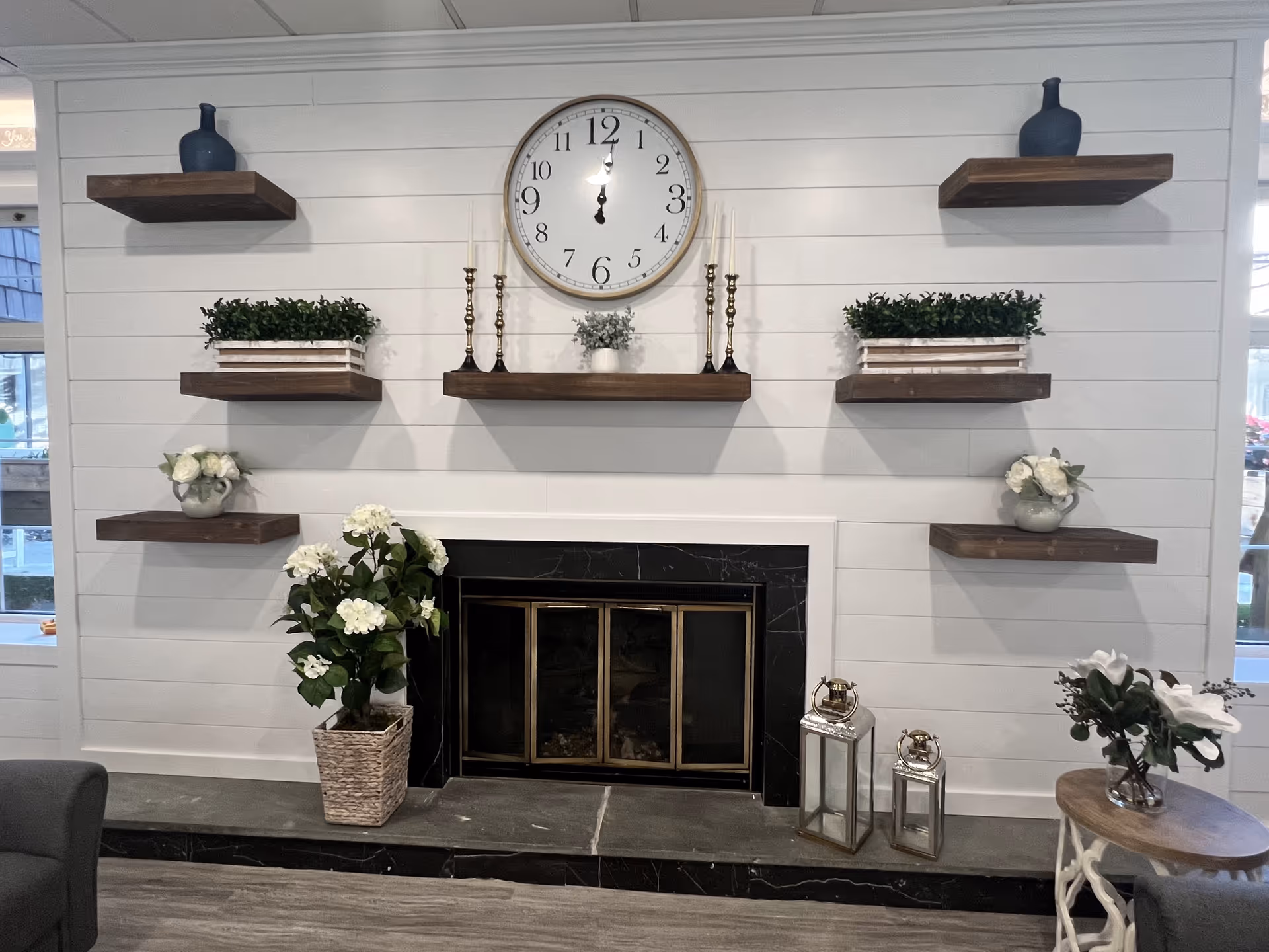 A cozy living room area featuring a black fireplace with a brass frame, surrounded by six wooden floating shelves mounted on a white shiplap wall. The shelves hold decorative items including blue vases, green plants in white wooden boxes, white flowers in small pots, and brass candlesticks. Above the fireplace is a large round wall clock showing the time as 12:00. On the hearth, there are two silver lanterns and a potted plant with white flowers. A small round wooden table with white legs and a vase with white flowers is positioned to the right. Part of a gray armchair is visible on the left side of the image.