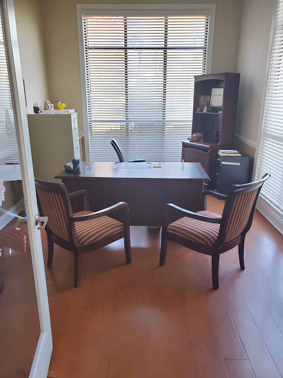 An office room with a wooden desk and two striped cushioned chairs facing it. Behind the desk is an office chair, a filing cabinet, and a wooden shelving unit. The room has large windows with blinds partially closed, allowing natural light to enter.