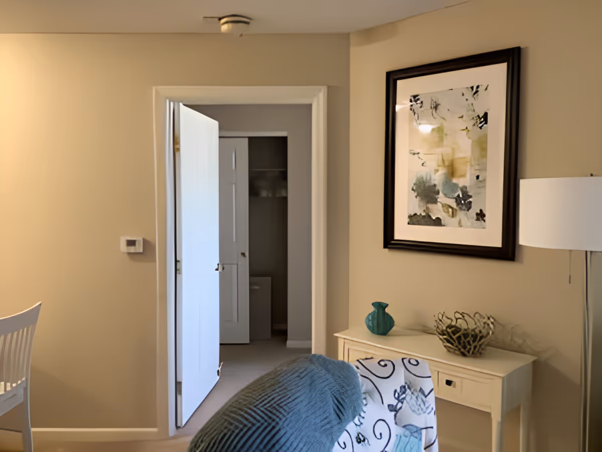 Interior view of a room with beige walls, a white door partially open leading to another room, a white chair on the left, a small white table with decorative items including a turquoise vase and a metal basket, a floor lamp with a white shade, and a framed abstract painting on the wall.