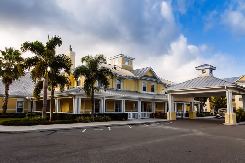 Exterior view of a senior living facility building with yellow walls, white trim, and a metal roof. The building features a covered entrance with columns and several palm trees in front. The sky is partly cloudy.