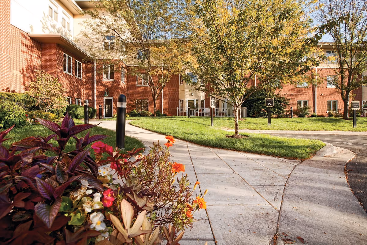 Outdoor view of Claridge Court showing a brick building with multiple windows, a paved walkway, green grass, trees, and colorful flowers in the foreground under a clear sky.