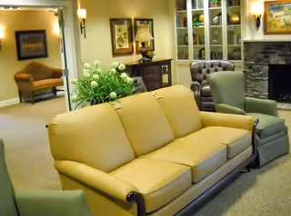 A cozy living room area with a yellow sofa, two green armchairs, and a brown leather armchair arranged around a stone fireplace. There is a wooden cabinet with a lamp and decorative items, a large plant, and framed artwork on the walls.