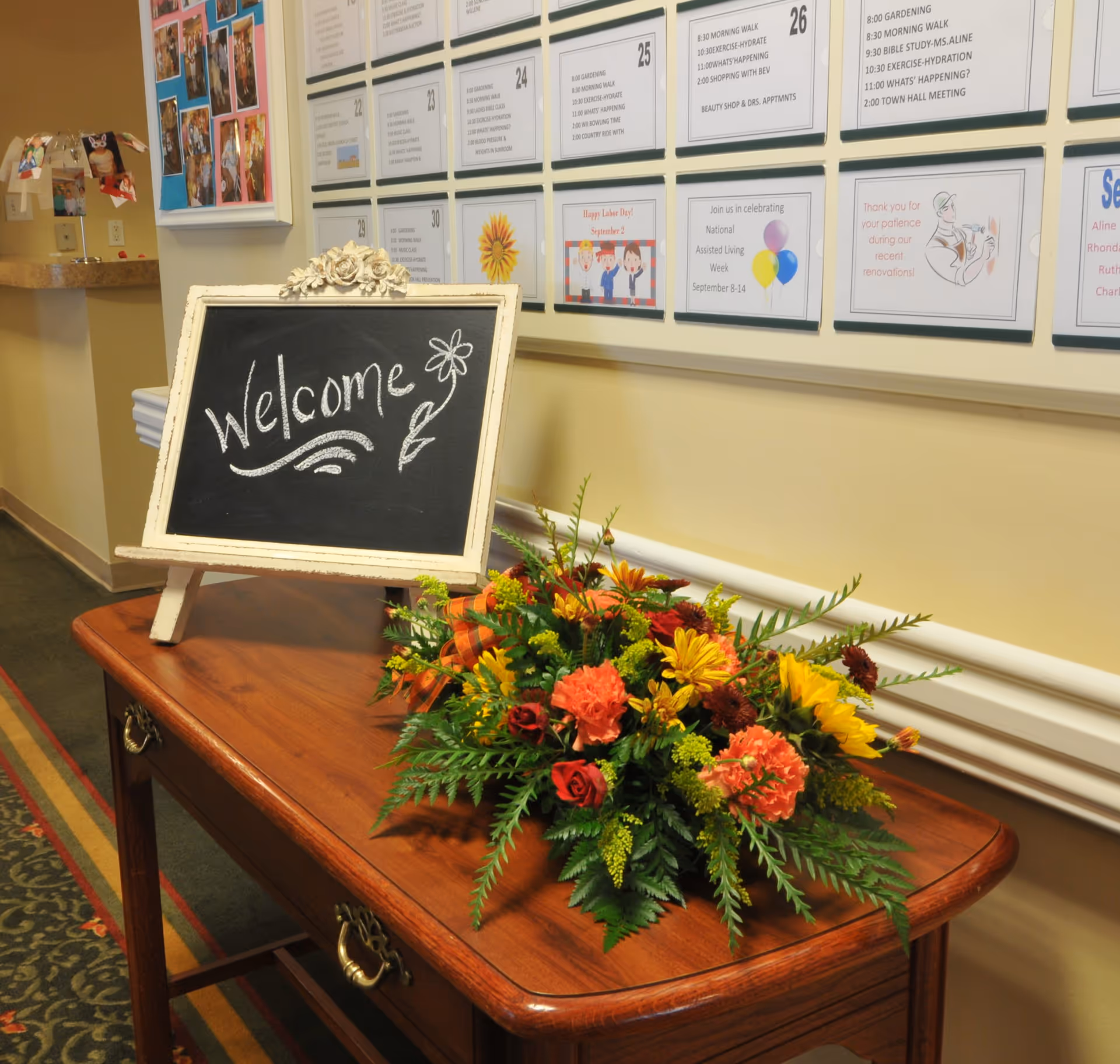 A wooden table with a floral arrangement featuring orange, yellow, and red flowers and green foliage. On the table is a small chalkboard sign with the word 'Welcome' and a flower drawing. Behind the table is a wall with various notices and schedules displayed in frames.