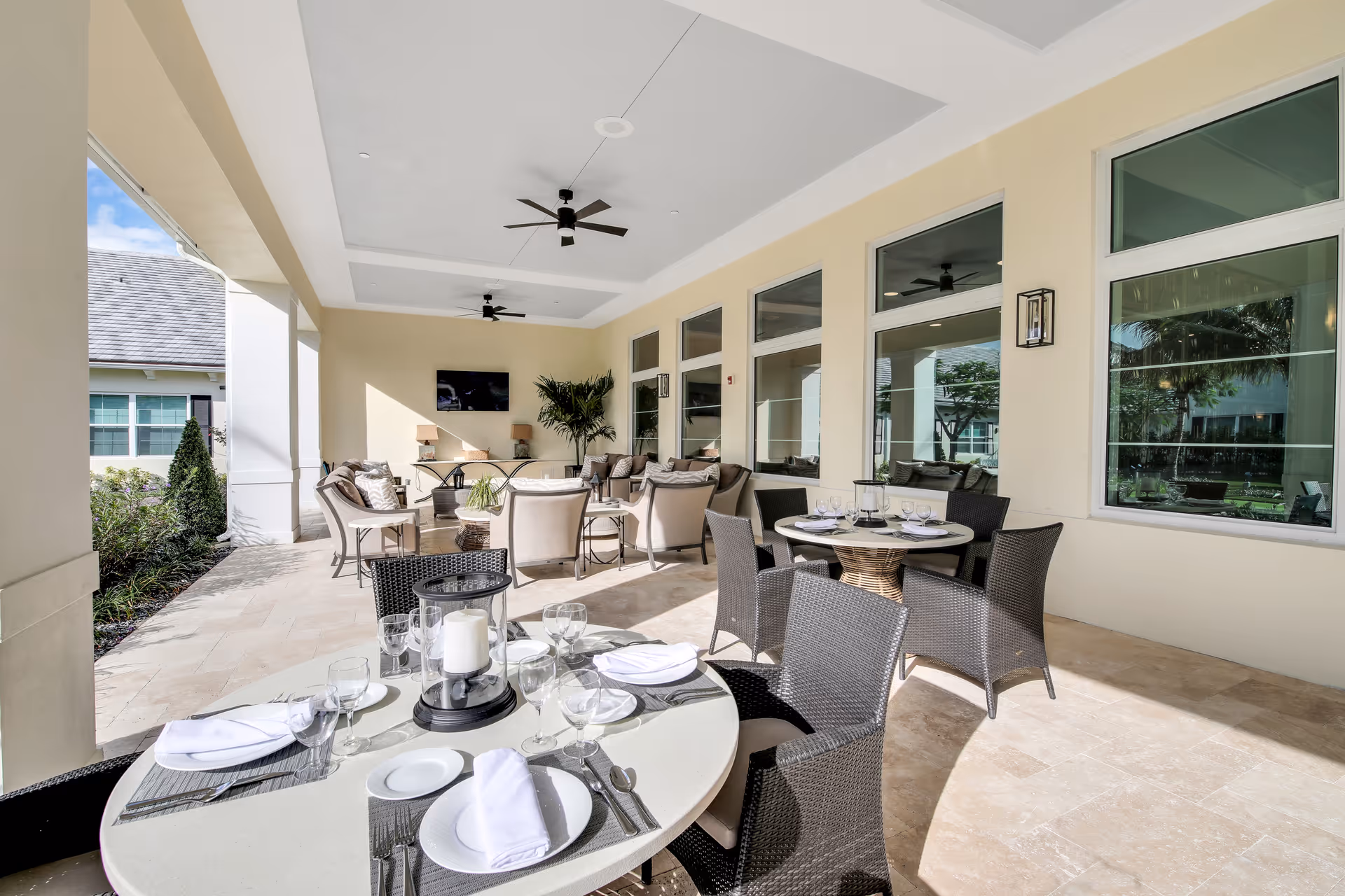 Covered outdoor patio area with dining tables set with plates, glasses, and napkins. Comfortable seating with cushioned chairs and sofas, ceiling fans, a mounted TV, and large windows looking into the building. The space is bright and airy with beige walls and tiled flooring.