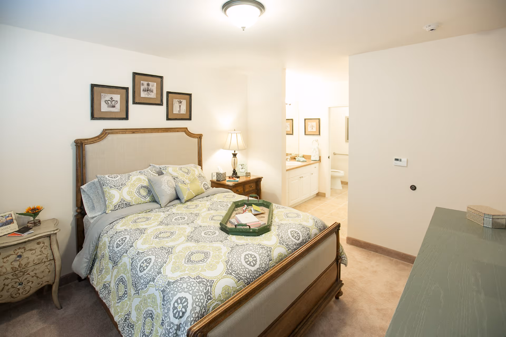 A cozy bedroom with a wooden bed frame and a patterned bedspread in shades of green, gray, and white. There are three framed pictures above the headboard. A nightstand with a lamp and decorative items is beside the bed. In the background, an open doorway leads to a bathroom with a sink, mirror, and toilet visible. The room has light-colored walls and carpeted flooring.