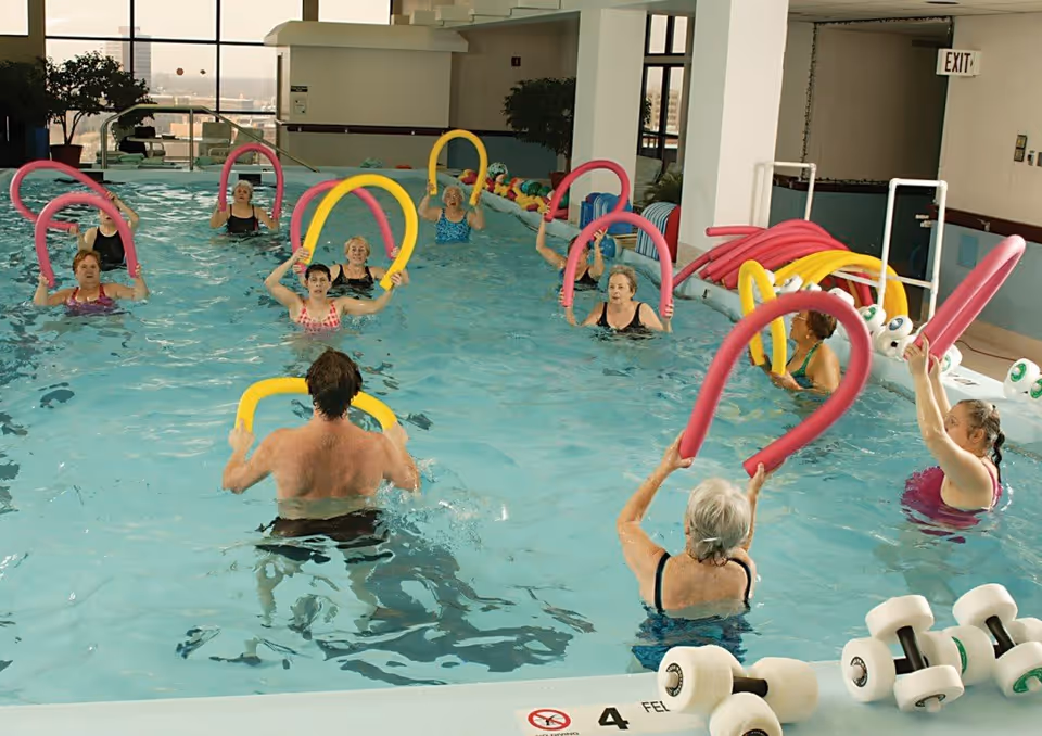 A group of senior adults participating in a water exercise class in an indoor swimming pool, each holding colorful foam pool noodles. The pool area has large windows and some pool equipment like dumbbells and extra noodles are visible on the side.