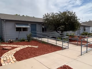 Exterior view of a single-story building with gray siding and a sloped roof. There is a concrete walkway with metal handrails leading to a blue door. The landscaping includes red mulch, a small tree, and a curved stone pathway.