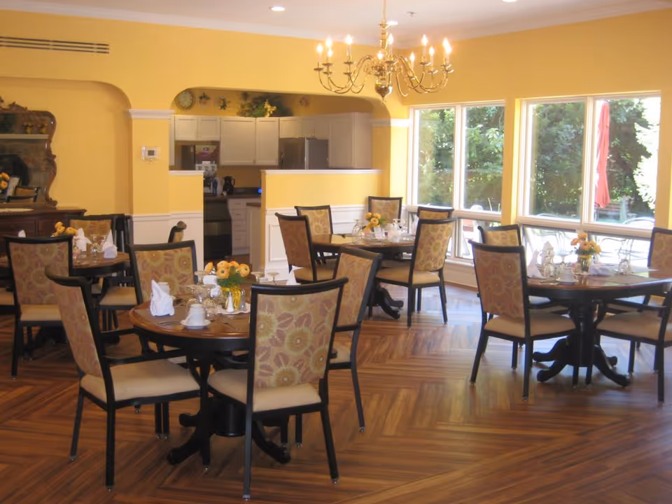 Bright dining room with multiple round tables set for meals, upholstered chairs, a chandelier, and large windows overlooking greenery.