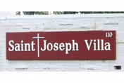 Maroon sign mounted on a white brick wall reading 'Saint Joseph Villa' with a cross forming the 't' and the number 110.