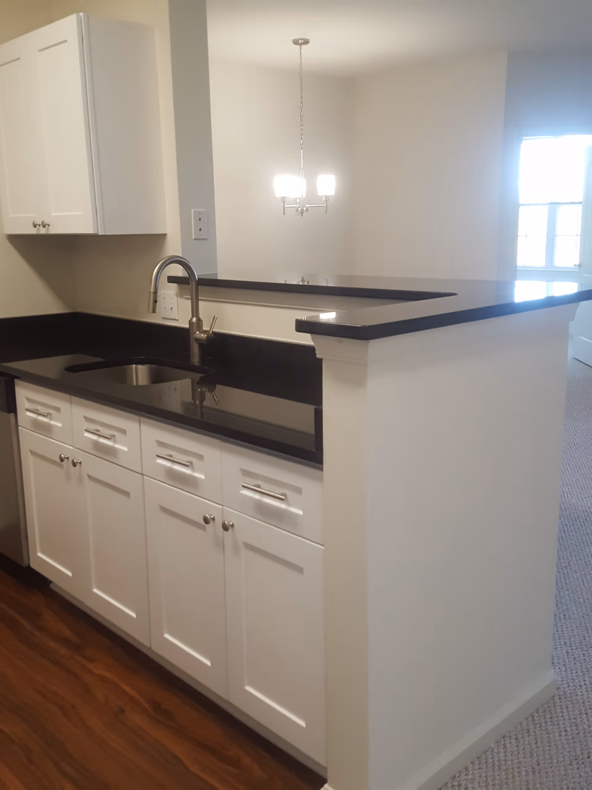 Interior view of a kitchen area with white cabinets, a black countertop, a stainless steel sink with a modern faucet, and a small bar counter. In the background, there is a hanging light fixture and a window letting in natural light.