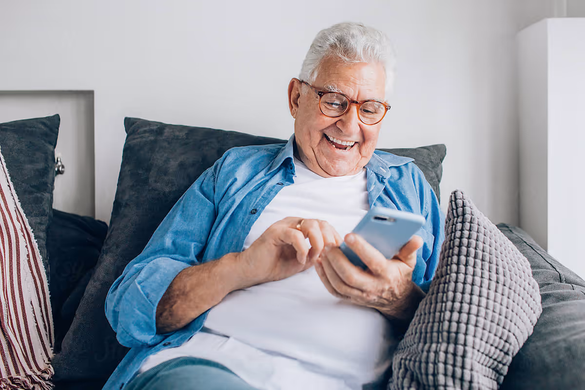 An elderly man with white hair and glasses sitting on a couch, smiling and using a smartphone. He is wearing a white t-shirt and a blue button-up shirt, surrounded by several cushions.
