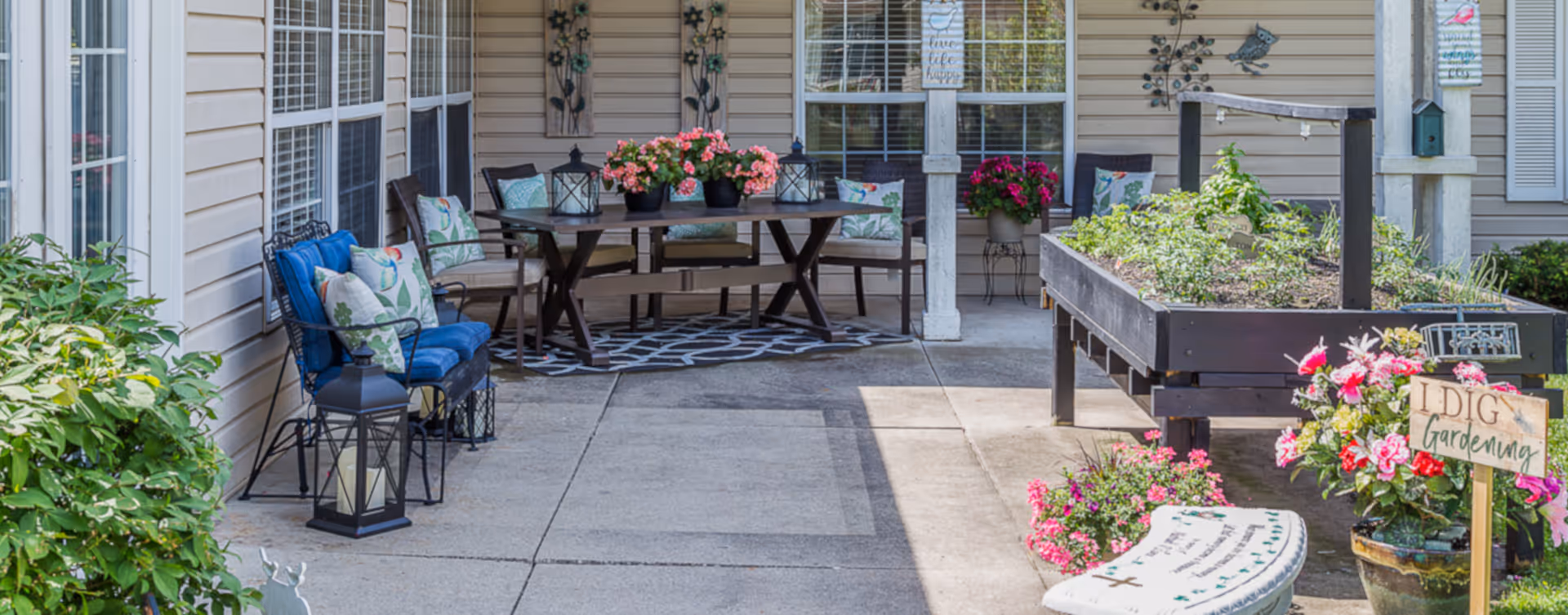 Outdoor patio area at Bickford of Urbandale featuring a seating arrangement with cushioned chairs and a table adorned with flower pots and lanterns. Raised garden beds with plants and flowers are visible, along with decorative signs and wall art on the building's exterior.