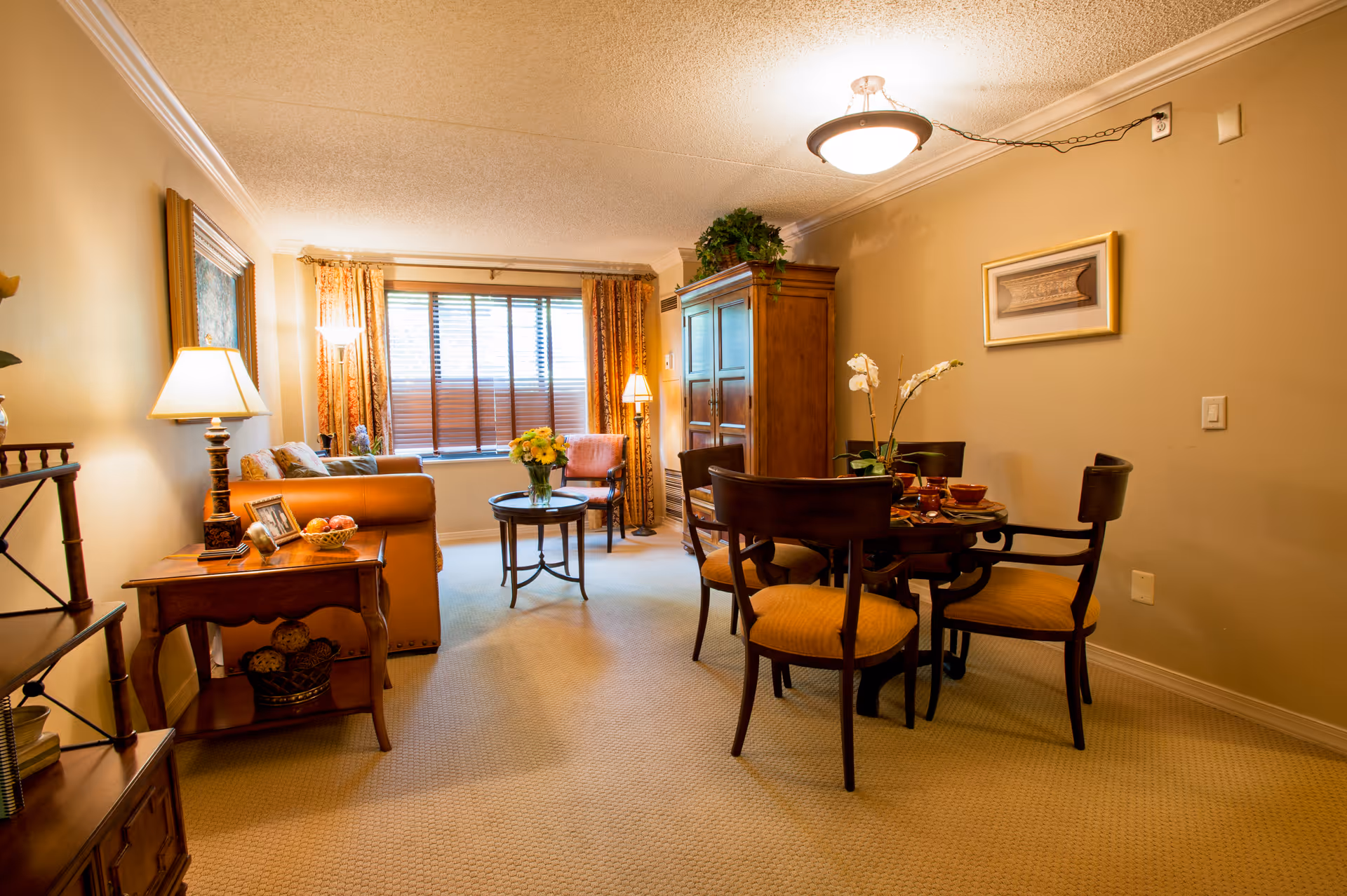 A cozy living and dining area in a senior living facility with beige walls and carpet. The living area features a brown leather sofa, a side table with a lamp and decorative items, a small round coffee table with a flower vase, and a window with curtains and blinds. The dining area has a round wooden table with four chairs, set with bowls and a flower centerpiece. A wooden cabinet and framed artwork are on the walls, and ceiling lighting illuminates the room.