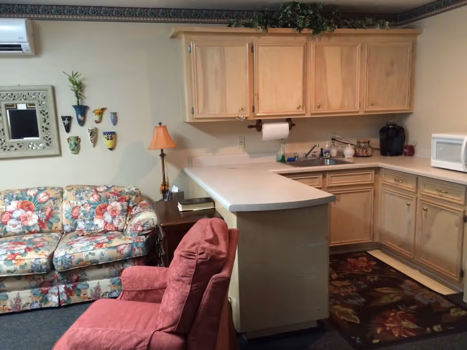 A cozy living area with a floral patterned couch and a red armchair next to a small wooden side table with a lamp and books. Adjacent to the seating area is a kitchen with light wood cabinets, a countertop with a paper towel holder, a sink, a coffee maker, and a microwave. The walls are decorated with small colorful wall planters and a mirror, and there is a floral rug on the floor in front of the kitchen cabinets.