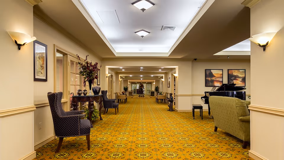 A long, well-lit hallway in a senior living facility with patterned yellow carpet, beige walls, and ceiling lights. The hallway is furnished with several chairs, tables with decorative vases and flowers, and a green sofa near a black grand piano. Framed artwork hangs on the walls.