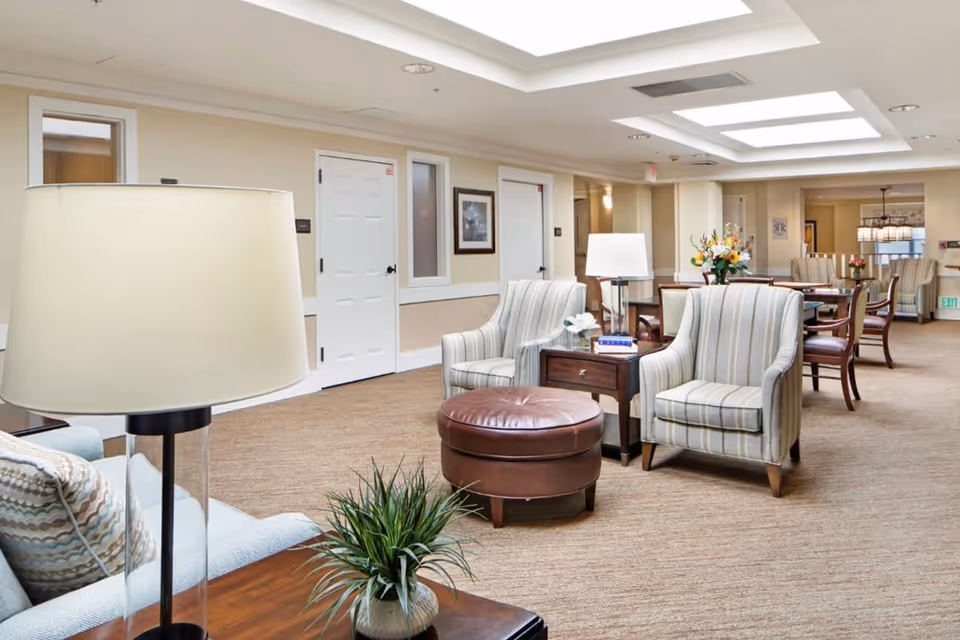 A spacious and well-lit senior living common area with beige walls and carpet. The room features comfortable striped armchairs, a round leather ottoman, wooden side tables with lamps, and a small potted plant. In the background, there are dining tables and chairs, floral arrangements, and framed artwork on the walls.