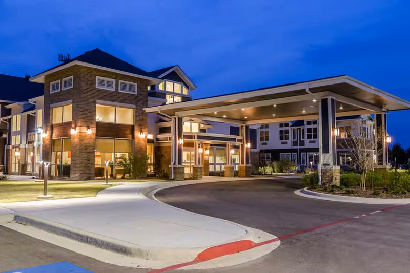 Exterior view of Ignite Medical Resort Fort Worth at dusk, showing a modern building with large windows, warm lighting, a covered entrance driveway, and landscaped surroundings.
