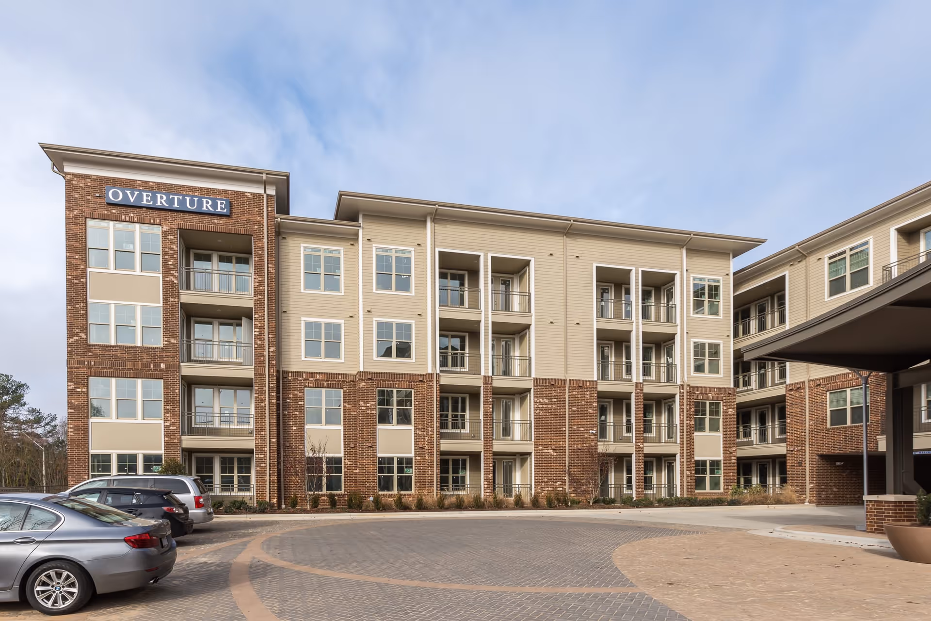 Exterior view of a modern multi-story apartment building with brick and beige siding, balconies, and multiple windows. Several cars are parked in front of the building under a partly cloudy sky. The building has a sign that reads 'OVERTURE'.