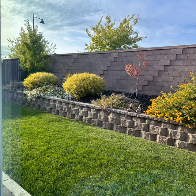 A landscaped outdoor garden area with a green lawn in the foreground, a raised stone flower bed containing various shrubs and flowering plants, and a brown shingled fence in the background under a partly cloudy sky.