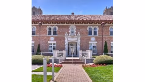 Front exterior view of a large brick building with a tiled roof, symmetrical windows, and a central entrance with steps and railings. The building is surrounded by neatly trimmed bushes, green grass, and a paved walkway leading to the entrance under a clear blue sky.