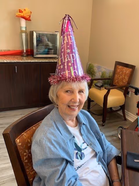 An elderly woman wearing a purple birthday party hat with 'Happy Birthday' written on it, sitting in a chair in a room with light-colored walls and wooden flooring. Behind her is a cabinet with a vase holding a large artificial flower and a small appliance. There are two chairs and a walker in the background.