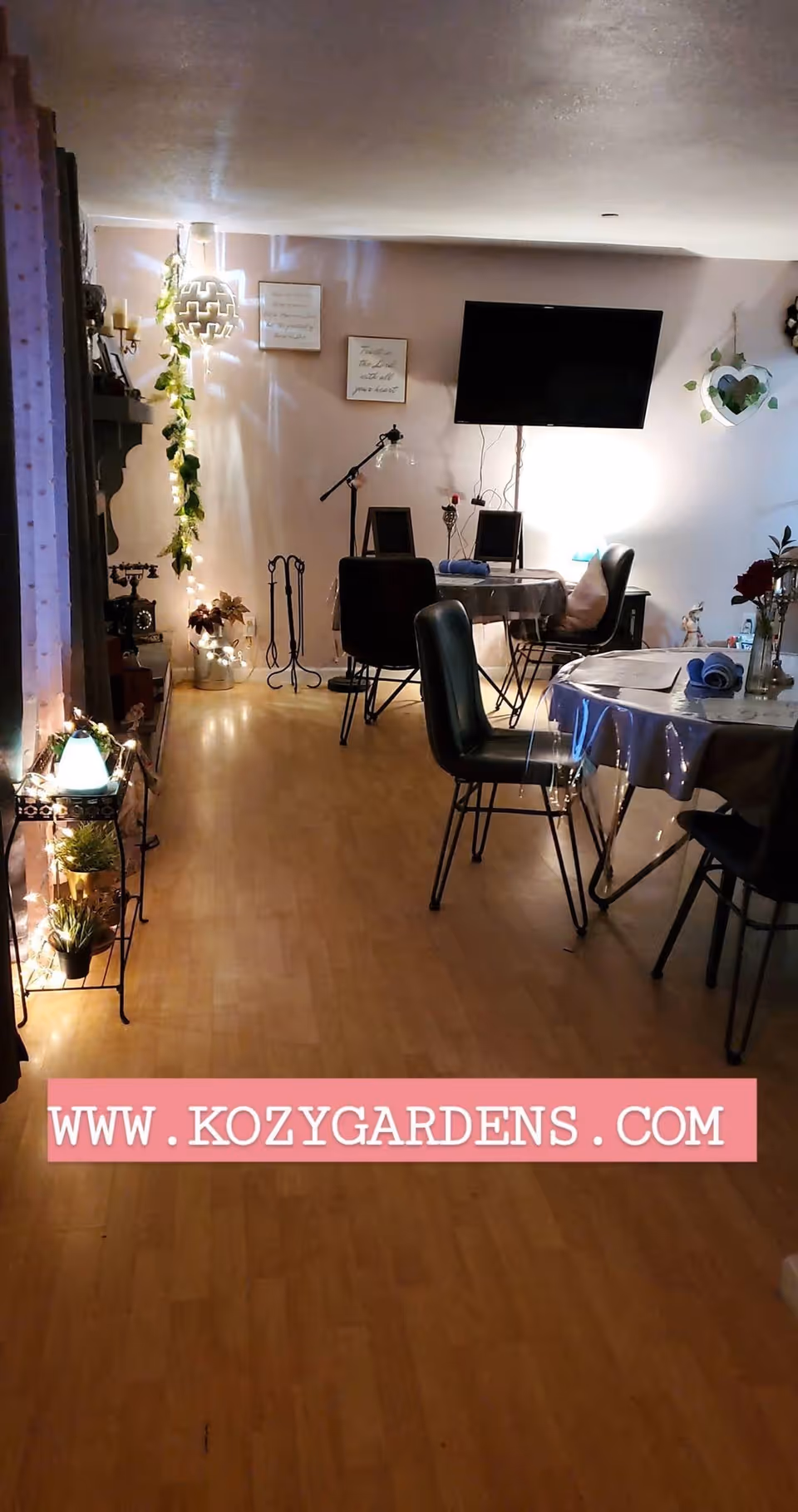 Interior view of a cozy assisted living common area with wooden flooring, a wall-mounted TV, several tables and chairs, decorative plants, and soft lighting. The room has a warm and inviting atmosphere with wall decorations and a lamp on a small table.