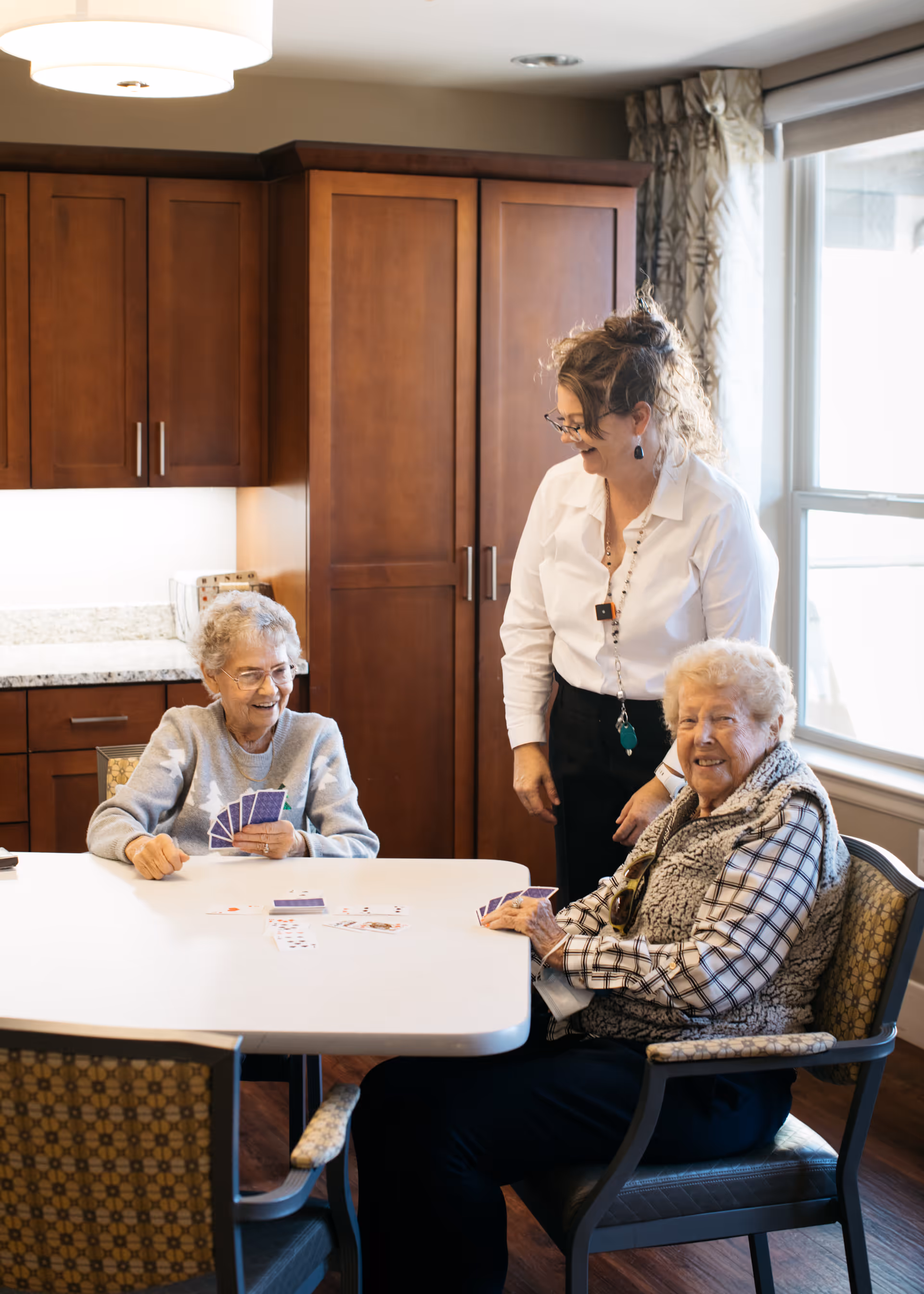 Two elderly women seated at a table playing cards while a staff member stands smiling in a dining area with wooden cabinets and a large window.