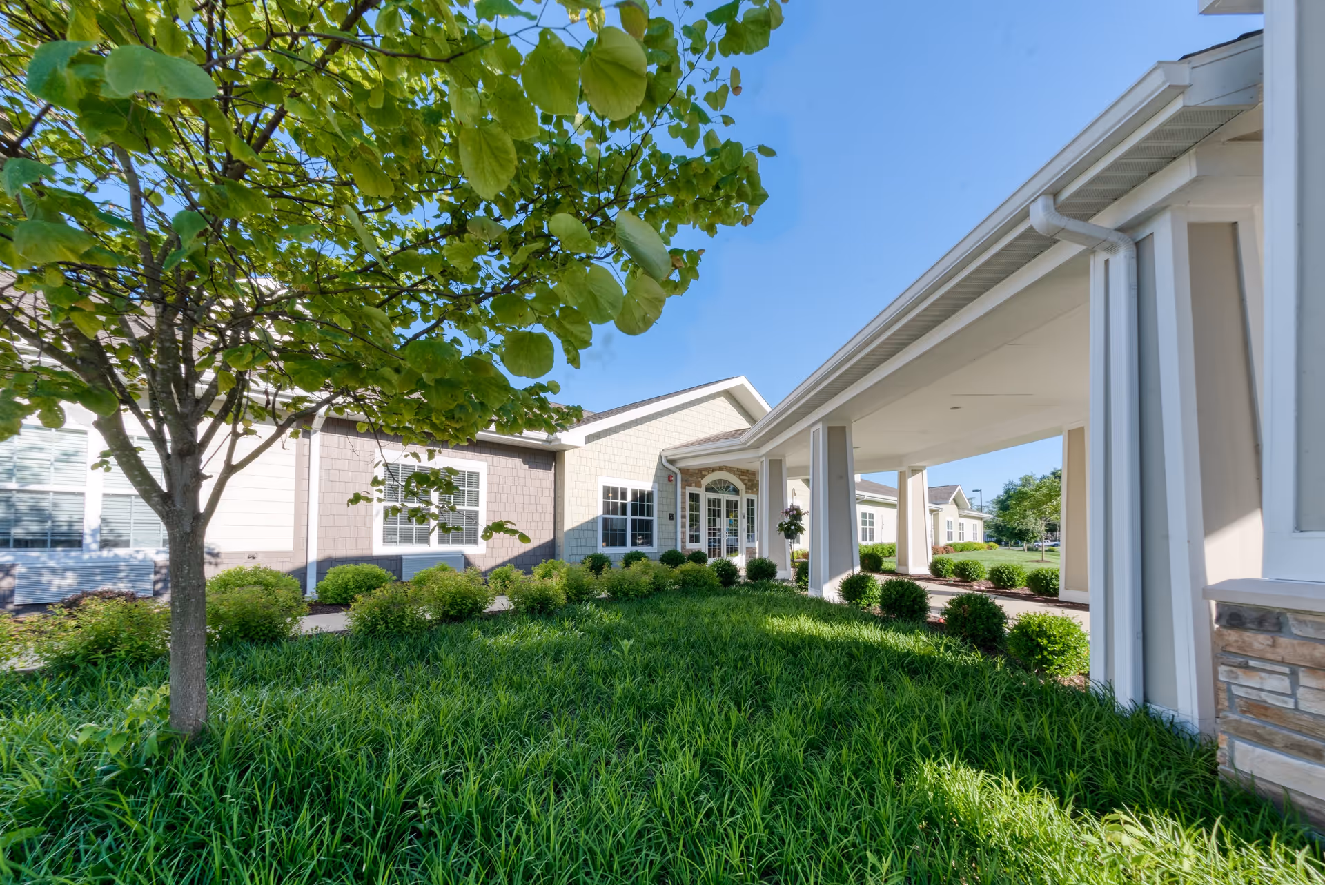 Exterior view of a senior living facility with a covered entrance, green shrubs, a tree, and a clear blue sky.