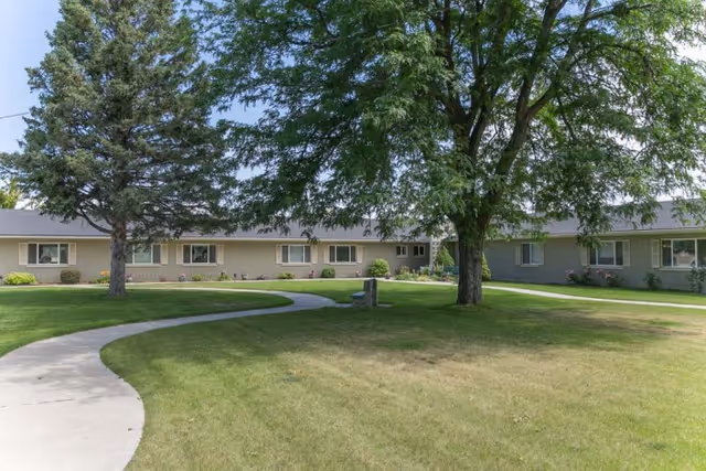 Single-story senior living building facing a grassy courtyard with winding concrete paths, large shade trees, and a bench.