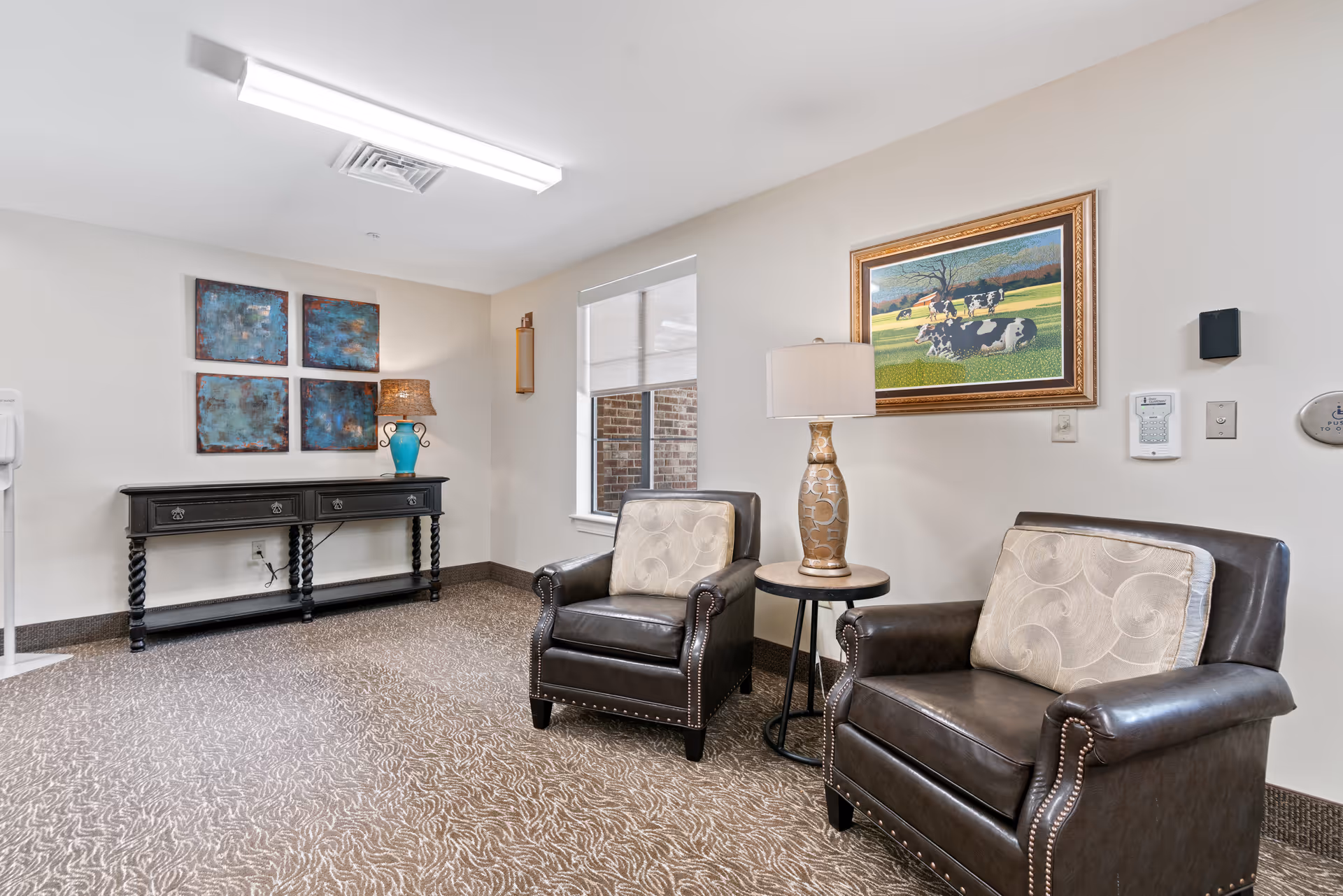 Two leather armchairs with pillows and a side table and lamp in a well-lit lounge area with artwork and a console table along the wall.