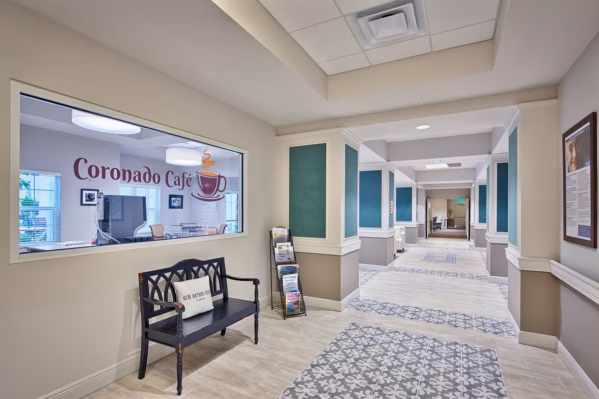 Interior hallway of a senior living facility with patterned tile flooring and beige walls. On the left side, there is a window with the sign 'Coronado Café' and a graphic of a steaming coffee cup. Below the window is a black wooden bench with a cushion. A brochure stand is next to the bench. The hallway extends into the distance with teal accent panels on the walls and framed pictures on the right wall.