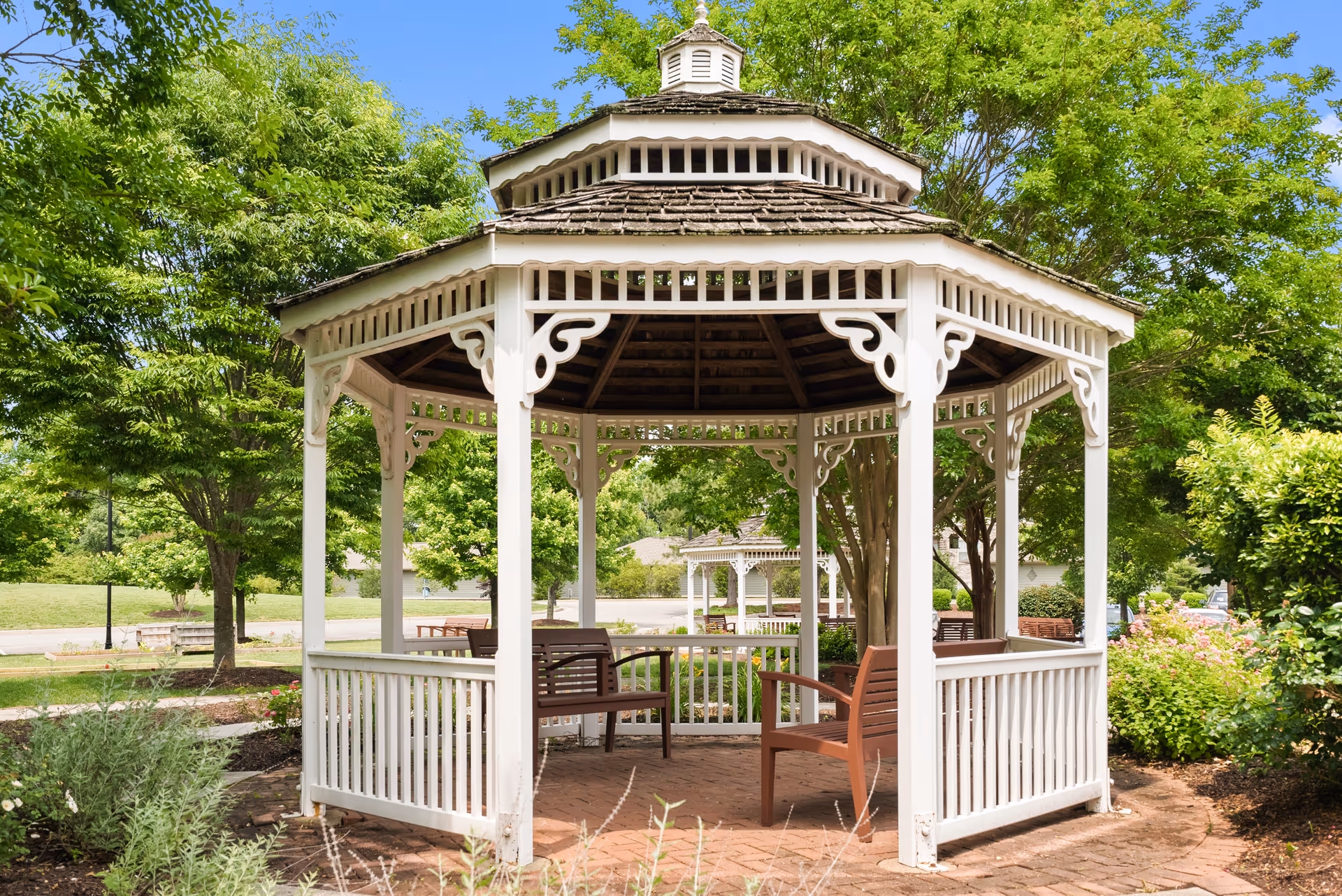 A white wooden gazebo with decorative trim and a shingled roof situated in a garden area with green trees, shrubs, and a clear blue sky in the background. There are wooden benches inside the gazebo and a paved brick floor.