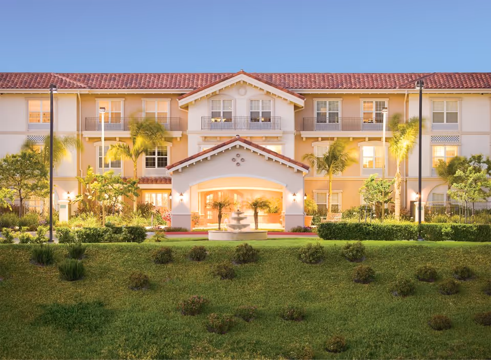 Exterior view of a three-story senior living facility building with a red tile roof, multiple windows, and balconies. The entrance features a covered porch with a fountain in front, surrounded by well-maintained landscaping including palm trees, shrubs, and grass under a clear blue sky.