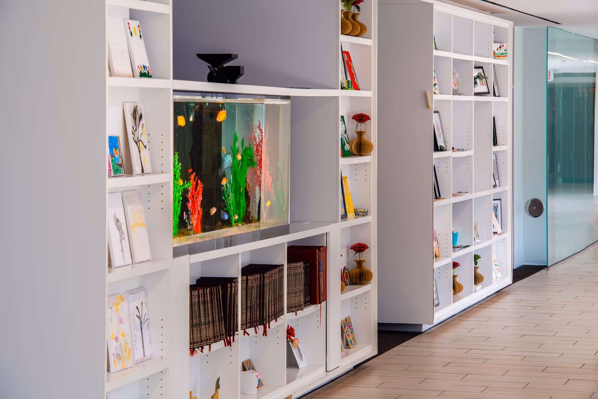 A modern interior hallway featuring white shelving units filled with books, framed pictures, small decorative items, and a built-in aquarium with colorful fish and aquatic plants. The floor is tiled, and a glass door is visible at the end of the hallway.